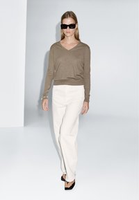 Beige Strickpullover mit langen Ärmeln und V-Ausschnitt, kombiniert mit weißen weit geschnittenen Hosen und schwarzen Sandalen mit eckiger Zehenpartie. Minimalistisches Design, glatte Textur.