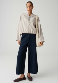 Frau trägt eine beige Reißverschlussjacke, ein gestreiftes Hemd, weite marineblaue Hose, braune Ballerinas und hat eine beige Tragetasche über der Schulter.