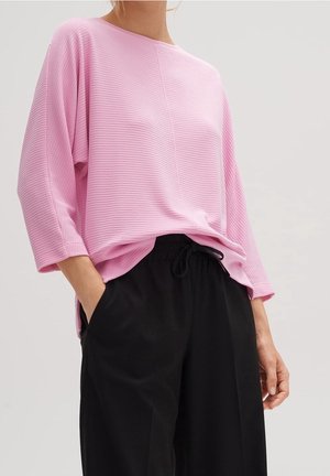 Un mannequin porte un haut rose ample en maille côtelée à manches trois-quarts et un pantalon noir à cordon de serrage, et pose avec une main dans une poche.