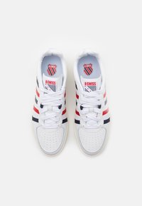 Bílé sportovní boty s červenými a námořnickými pruhy, kulatou špičkou, perforovanými detaily a logem K-Swiss na jazyku. Materiály z textilu a gumy.