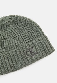 Bonnet tricoté en vert olive avec une texture côtelée. Présente une bordure repliée et un logo "CK" brodé en gris foncé sur le côté.
