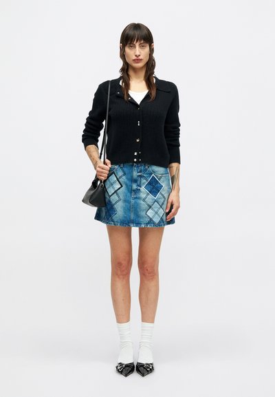 Vrouw in een zwarte gebreide cardigan, witte top, denim minirok met diamantenpatroon, witte sokken, zwarte puntige schoenen en met een zwarte handtas.