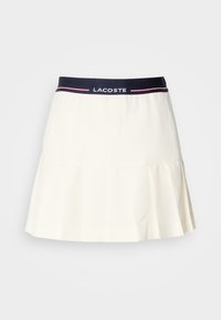 Vit plisserad kjol med midja i marinblått och en Lacoste-logotyp samt röda detaljer. Tillverkad av ett mjukt och slätt material.