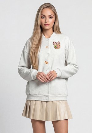 Ung kvinde med langt blondt hår har en hvid knap-sweatshirt med bjørne-lapper og en beige plisseret nederdel på, står med hænderne foldet.