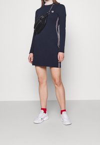 Marinblå långärmad klänning med vita och vinröda sidosträck, med rund hals. Bärs med en svart crossbody-väska och vita sneakers.