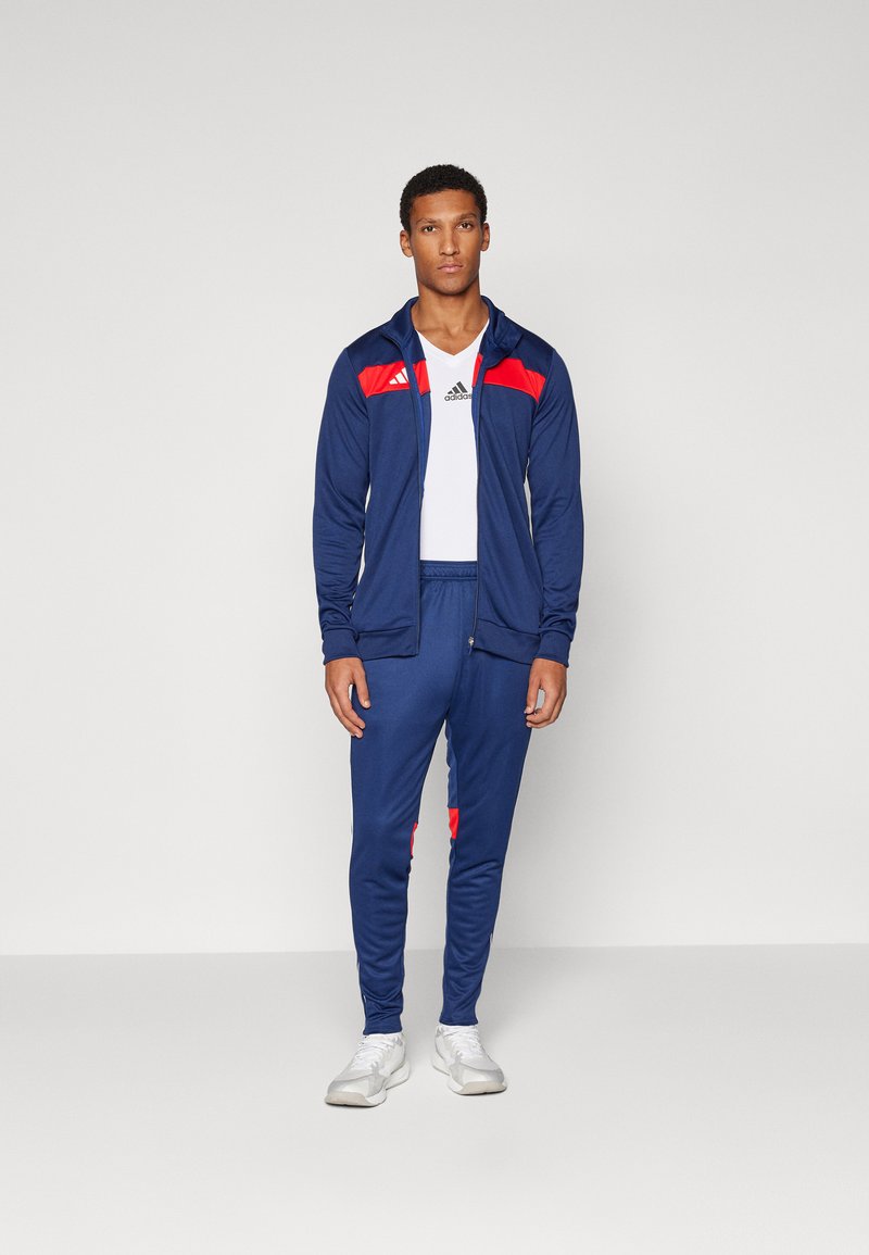 Chándal de color azul marino con acentos rojos, que incluye una chaqueta con capucha y cremallera y pantalones ajustados. Hecho de tejido transpirable, incluye el logo de Adidas.
