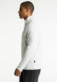Hvid zip-up fleece pullover med højt krave, lange ærmer og et logo patch på siden, parret med mørke jeans, vist i profil.