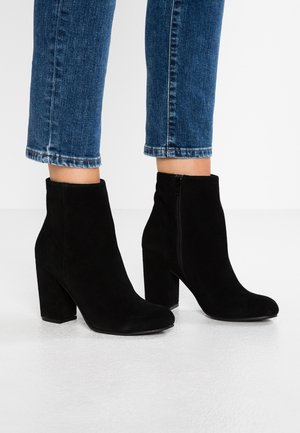 Bottes basses en daim noir avec un talon haut carré et une fermeture éclair latérale, associées à un jean bleu raccourci. Texture lisse, design simple.