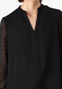 Nainen, jolla on yllään musta paita, jossa on halkio mandariinikaulus ja läpinäkyvät kuvioidut hihat, joissa on viivoja.