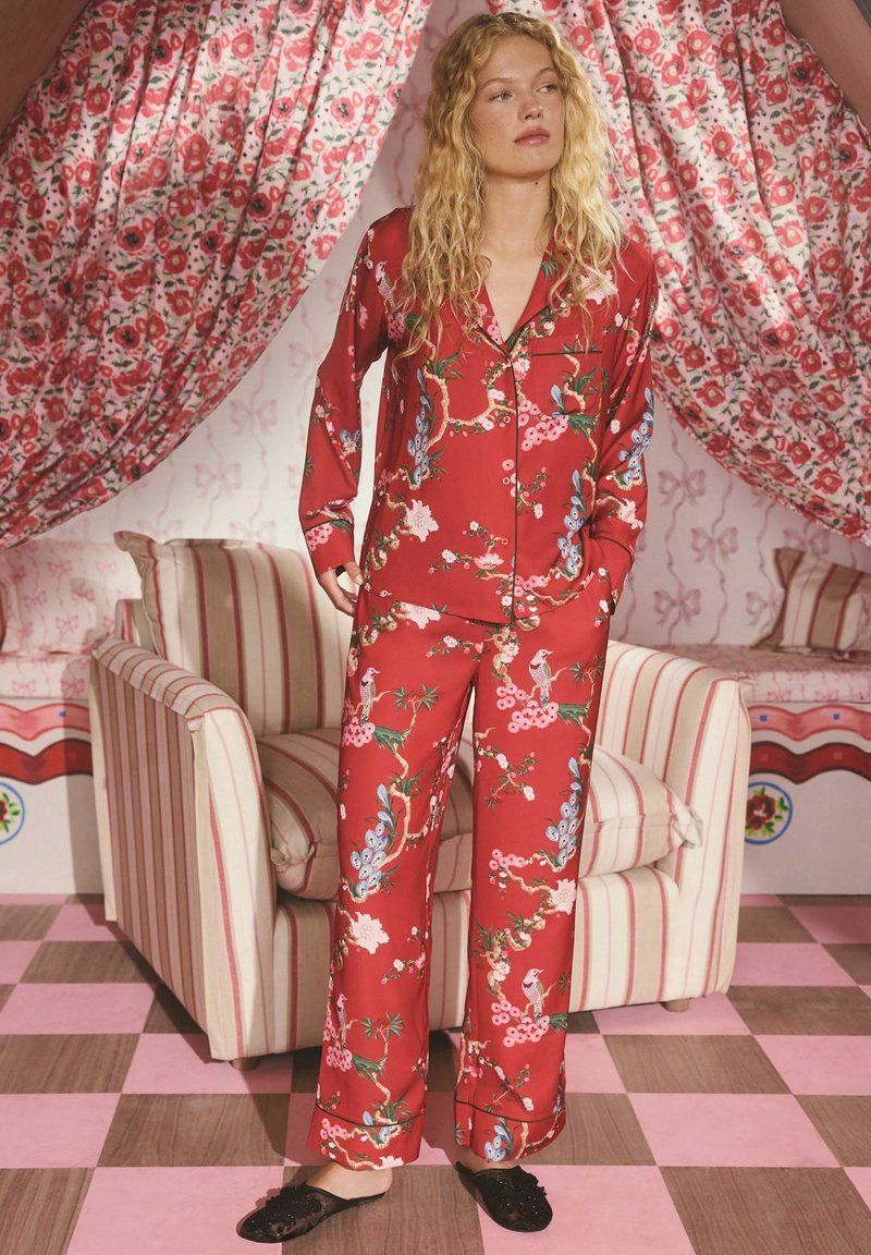 Set de pijamale din satin roșu cu imprimeu floral, cu mâneci lungi și pantaloni, având o croială lejeră și modele botanice detaliate. Pantofi negri slip-on.