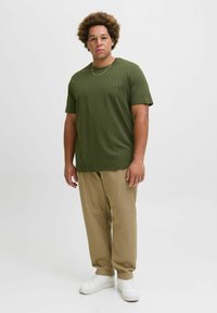 Khaki bukser kombinert med en mørkegrønn t-skjorte med en subtil logo, komplementert med hvite joggesko og et sølvkjede.