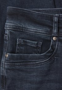 Tummansiniset farkut lähikuvassa, jossa näkyvät etutasku, pieni kolikoille tarkoitettu tasku, vyölenkki ja metalliset niitit teksturoidulla kankaalla.