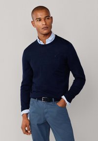 Marineblauwe crewneck trui gemaakt van lichtgewicht stof, over een lichtblauw overhemd met kraag gedragen. Slank gesneden blauwe broek en een zwarte riem maken de look compleet.