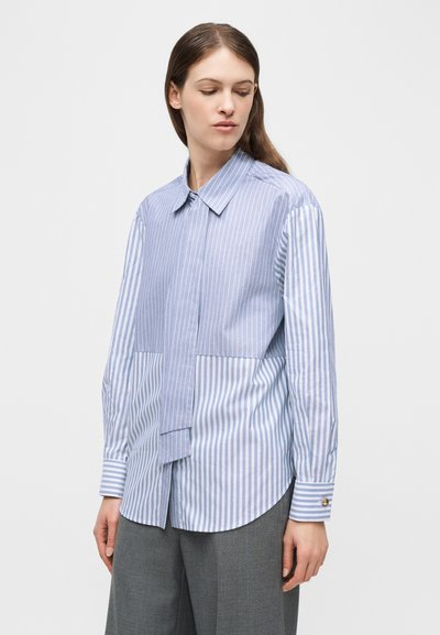 Γυναίκα που φοράει μπλε και λευκό κάθετο ριγέ πουκάμισο με κουμπιά και μακριά μανίκια και γκρι παντελόνια, κοιτάζοντας προς τα κάτω αριστερά της.