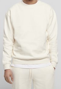 Felpa di colore crema chiaro, realizzata in morbido cotone, presenta un collo a girocollo e polsini a coste, con una vestibilità rilassata e una sottile trama. Indossata sopra una t-shirt bianca.