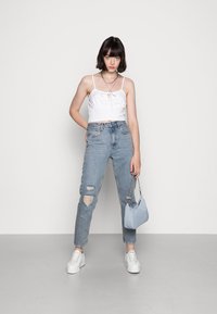Top cropped branco com um detalhe de laço, jeans claros desbotados, tênis brancos e uma pequena bolsa azul. Fundo branco liso.