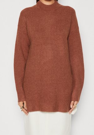 Vrouw draagt een roestkleurige geribbelde gebreide trui met lange mouwen en een hoge hals, gecombineerd met een witte rok.