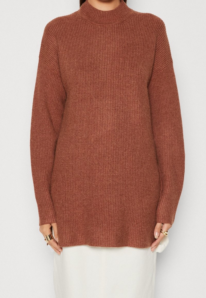 Femme portant un pull en maille côtelée couleur rouille avec des manches longues et un col montant, associé à une jupe blanche.