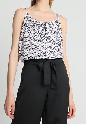 Kvinde iført en ærmeløs hvid top med sorte prikker og højtaljede sorte bukser bundet med en sløjfe i taljen.