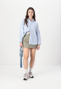 Camicia azzurra a righe bianche con bottoni, gonna verde oliva, sneakers pastello e borsa blu con tracolla. Sfondo bianco.