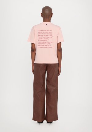 Persoon draagt een lichtroze T-shirt met rode tekst en bruine wijdvallende broek, staand met de rug naar de camera tegen een effen witte achtergrond.