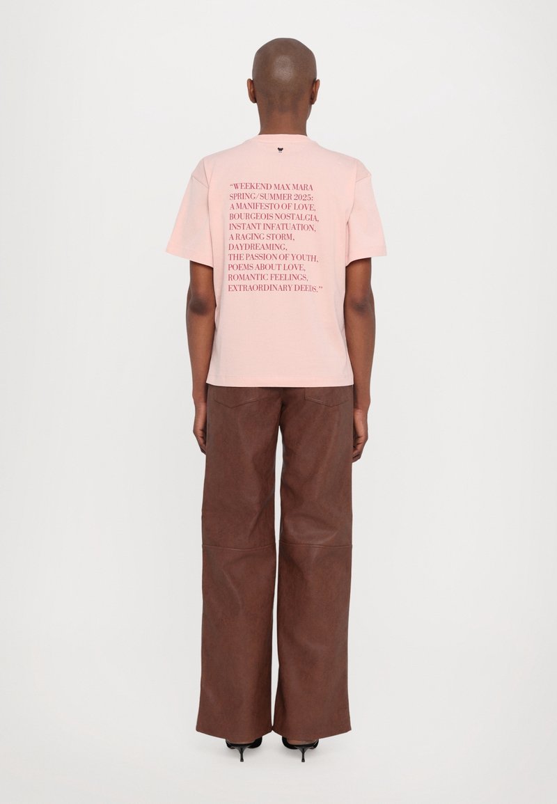 Personne portant un t-shirt rose clair avec un texte rouge et un pantalon marron à jambes larges, debout, dos à la caméra, sur un fond blanc uni.