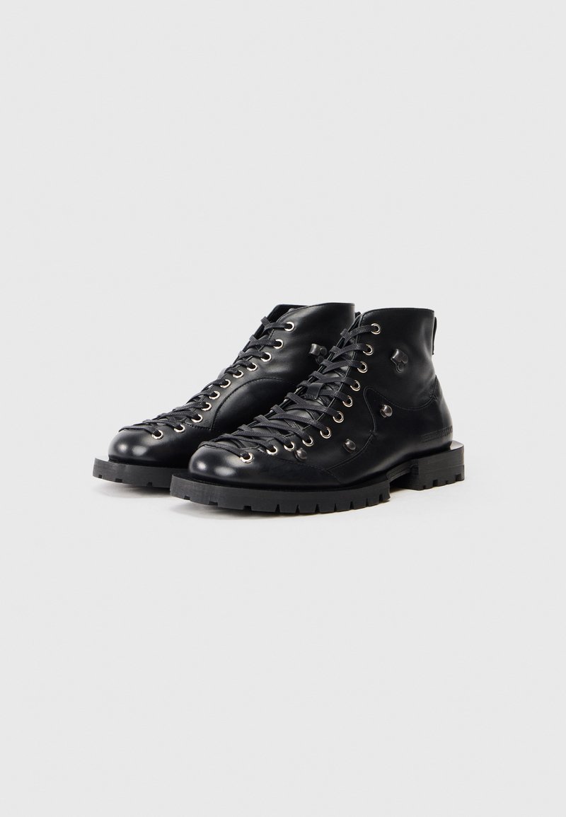 Ein Paar schwarze Leder-Stiefeletten mit Schnürung, metallenen Ösen und dicken Gummi-Sohlen auf einem weißen Hintergrund.