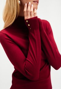 Kobieta w czerwonym swetrze z długim rękawem i golfem, z złotymi guzikami na mankietach, dotyka twarzy jedną ręką.