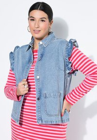 Spijkervest met ruches op de schouders, lichtblauw van kleur, voorzakken en knoopsluiting, gedragen over een rood gestreept topje.