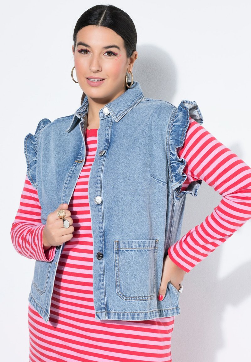 Spijkervest met ruches op de schouders, lichtblauw van kleur, voorzakken en knoopsluiting, gedragen over een rood gestreept topje.