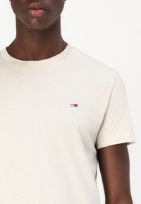 Beige bomull T-skjorte med korte ermer, rund hals, med en liten brodert logo i rød, hvit og blå på venstre bryst.