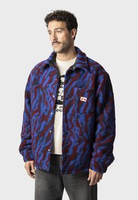 Fleece bunda v modrej a bordovej farbe s vlnitým vzorom. Má golierový výstrih, predné zapínanie na gombíky a náprsné vrecko s logom.