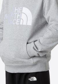 Hanorac gri cu glugă, cu un logo mare brodat alb pe piept, manșete striate, buzunar frontal și croială lejeră. Pantaloni negri cu logo vizibil.