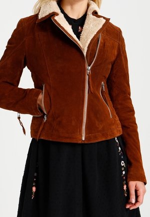Mujer con chaqueta marrón de ante con cuello de piel de oveja y cremalleras plateadas sobre una falda negra con bordados florales.