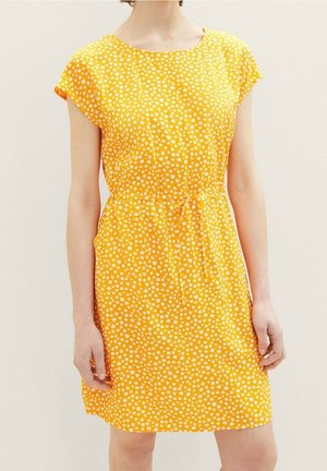 Vrouw draagt een geel knielang jurkje met kleine witte bloemenprint en een strikceintuur, staand tegen een lichte achtergrond.