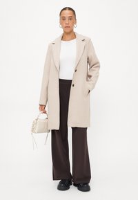 Beige wollen blendjas met notch-lapels, enkelbreasted ontwerp, twee voorkruimelpockets, gecombineerd met donkerbruine wijde broek en zwarte sneakers.