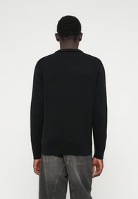 Vyras matomas iš nugaros, dėvintis juodą ilgo rankovės megztinį ir nusidėvėjusius juodus džinsus prieš paprastą baltą foną.