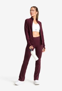 Vinröd träningsklädesset som innehåller en figurnära zip-up jacka och högmidjade byxor, tillsammans med en vit sport-bh och en vattenflaska.