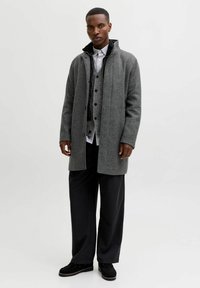 Grijze wollen mix jas met een hoge kraag, gecombineerd met een grijze vest met knopen en een zwart shirt. Losse zwarte broeken en zwarte schoenen maken de look compleet.