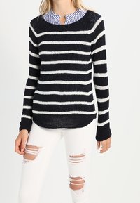 Jersey a rayas en azul marino y blanco con un tejido texturizado, cuello redondo y mangas largas, combinado con unos vaqueros blancos desgastados.