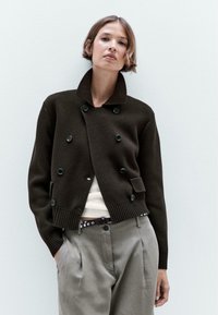 Brauner gestrickter Cardigan mit hohem Kragen, verziert mit grünen Knöpfen und Seitentaschen, kombiniert mit hellgrauen Hosen und einem Gürtel.