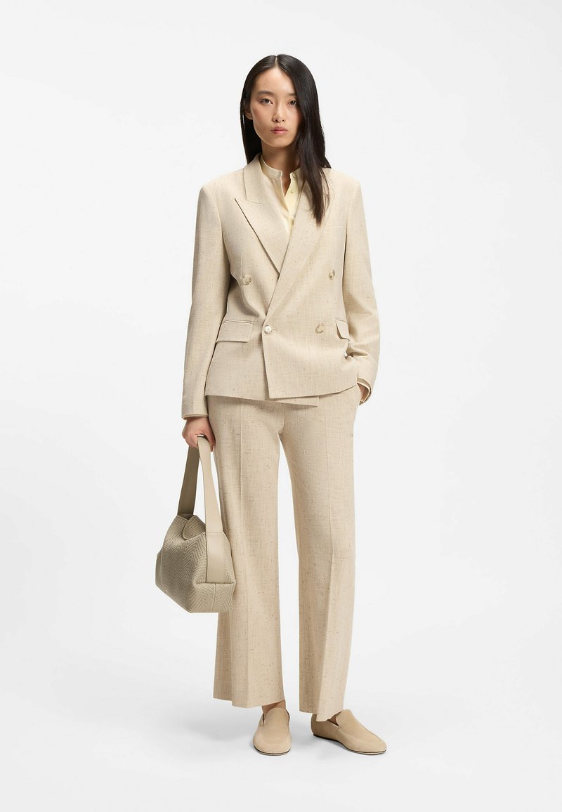 Femme debout, portant un blazer beige croisé, pantalon large assorti, chaussures beiges, tenant un sac à main beige texturé à ses côtés.