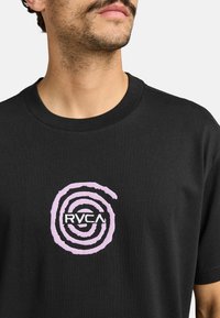 Musta puuvillapaita, jossa on violetti spiraalimalli ja "RVCA"-logo keskellä. Pyöreä kaula-aukko ja lyhyet hihat, sileä tekstuuri.