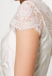 Haut blanc avec manches en dentelle transparente ornées de motifs floraux, bords festonnés et effilochage visible le long de l'ourlet des manches. Corps en tissu lisse.