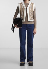 Person i brun ribbad tröja, beige broderad shearling-väst, blå jeans och plattformsskor som håller i en svart vadderad väska.