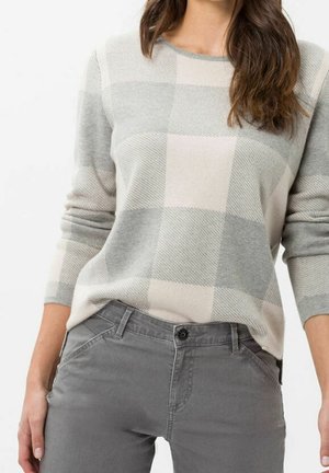 Femme portant un pull à manches longues à carreaux gris et beige, rentré dans un pantalon gris, debout les mains détendues le long du corps.