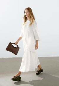 Top bianco a maglia grossa con maniche a tre quarti, abbinato a una gonna vaporizzante bianca. Una borsa a mano intrecciata marrone e sandali con piattaforma neri completano il look.