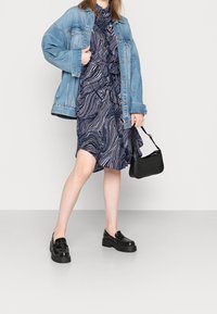 Farkkutakki tummansinisellä mekolla, jossa on valkoisia aaltokuvioita, yhdistettynä mustiin platform-loafereihin ja pieneen mustaan käsilaukkuun.