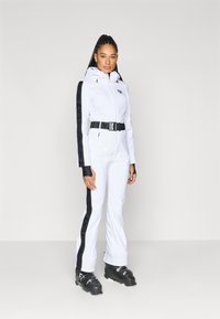 Λευκή στολή σκι με εφαρμοστή σχεδίαση, μαύρες λεπτομέρειες στα μανίκια και ζώνη. Διαθέτει κουκούλα και τσέπες με φερμουάρ. Φοράει μαύρα παπούτσια σκι.