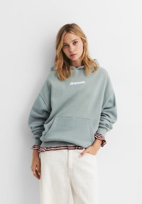 Jasnoniebieska, oversize bluza z kapturem z przednią kieszenią, z białym haftowanym napisem "Dramatic." Białe spodnie i w paski koszula pod spodem.